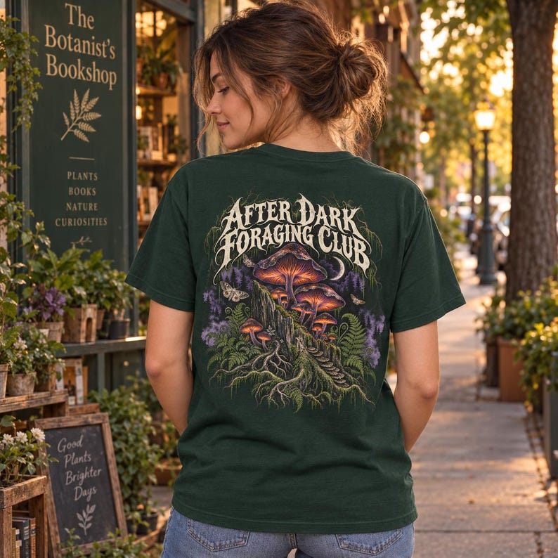 May include: A dark green t-shirt featuring a graphic on the back with the text After Dark Foraging Club. The illustration depicts glowing purple and orange mushrooms growing on a mossy log, surrounded by ferns, moths, and a crescent moon.