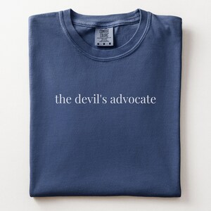 May include: A blue crew neck t-shirt featuring the text "the devil's advocate" printed in white across the center. The garment has a ribbed collar and a Comfort Colors brand label visible at the inner neckline, indicating a size medium.