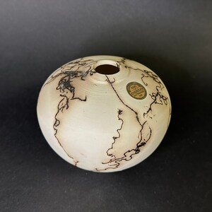 May include: A white ceramic vase with black markings. The vase is round and has a small opening at the top. It has a label that reads "HANDCRAFTED AMERICAN INDIAN ART".
