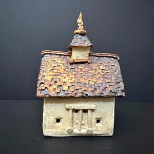 May include: A handcrafted ceramic house sculpture with a textured, multi-toned roof in shades of brown and gold. The house features a small tower and detailed facade with windows and a door. The sculpture is set against a dark background.