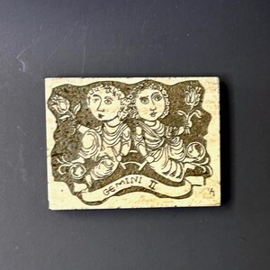 May include: A ceramic tile with a black and white illustration of the Gemini zodiac sign. The tile is beige with a brown background and the text "GEMINI II" is written on a banner.