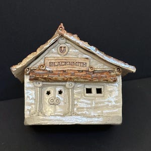 May include: A ceramic miniature blacksmith shop with a textured, off-white exterior. The roof is tan with a metallic sheen. The sign above the door reads "BLACKSMITH". The building has a door with star-shaped cutouts and two small windows.