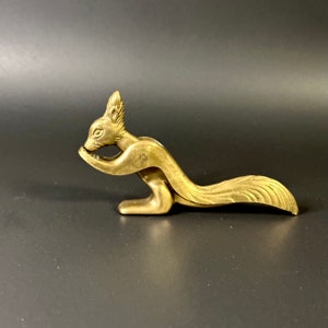 May include: A gold-colored squirrel nutcracker. The squirrel is depicted in a sitting position with its tail curled around its body. The nutcracker is made of metal and has a hinged jaw.