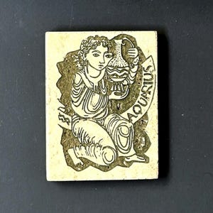 May include: A white tile with a black and white illustration of the astrological sign Aquarius. The illustration depicts a figure pouring water from a pitcher. The word "Aquarius" is written in a curved script around the figure.