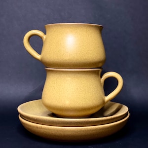 May include: Two stacked beige ceramic mugs with a matching saucer. The mugs have a rounded shape and a slightly flared lip.