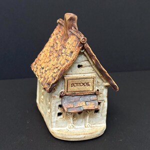 May include: A ceramic schoolhouse figurine with a textured, brown roof and a sign that reads "SCHOOL". The building is off-white with brown accents and small cut-out windows. The chimney is brown and the base is rounded.