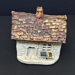May include: A miniature ceramic house with a textured brown roof and a chimney. The house has a white facade with decorative elements and a small window. The overall design is rustic and charming.