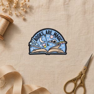 May include: A semi-circular embroidered patch featuring an open book with the text "BOOKS ARE MAGIC" arched above. The design includes a crescent moon, a skull, a feather, a mushroom, and stars on a light blue background with a black border.
