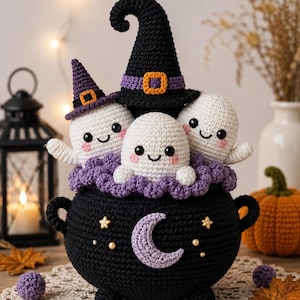 May include: A black crocheted cauldron featuring a light purple crescent moon and small star accents. Inside the cauldron are three white crocheted ghosts with pink cheeks, one wearing a tall black witch hat and another a smaller purple hat.
