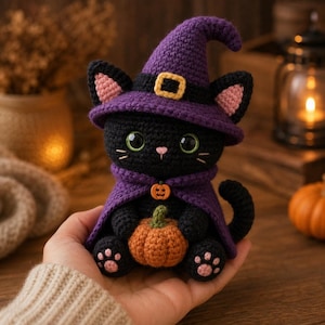 Könnte beinhalten: Eine gehäkelte schwarze Katzenfigur, die einen violetten Hexenhut mit gelber Schnalle und einen passenden violetten Umhang trägt. Die Katze hat große grüne Augen, rosa Innenohren und rosa Pfotenballen. Sie hält einen kleinen orangefarbenen Kürbis.