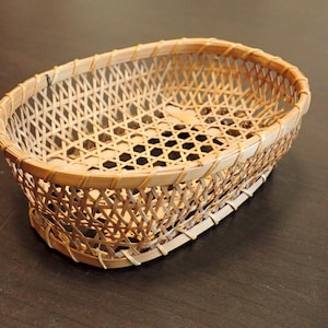 May include: Oval-shaped woven basket made of light brown bamboo. The basket features a lattice weave pattern with a reinforced rim. The base has a series of hexagonal openings. The basket is empty and sits on a dark surface.