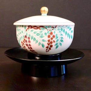May include: A white ceramic bowl with a lid, featuring a floral design in red, green, and gold. The bowl sits on a black, tiered stand. The lid has a gold knob.