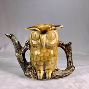 May include: A ceramic owl teapot with a brown and green glaze. The owl is perched on a branch with a spout on one side and a handle on the other.