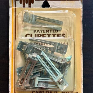May include: A package of Tip-Top patented clipettes hair clips. The package is a mix of tan, yellow, and brown with the words "Tip-Top" and "Patented Clipettes" printed on the front. The package contains a card of 12 silver metal hair clips.