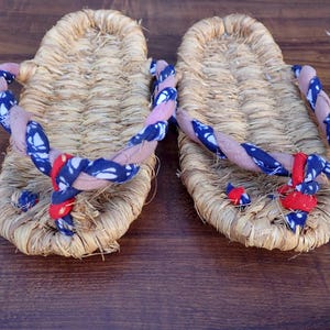 May include: A pair of traditional Japanese straw sandals, or Zori, with woven straw soles. The straps are a combination of pink, blue, and red fabric. The sandals are displayed on a dark wooden surface.