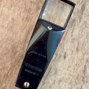 May include: A black handheld magnifying glass with a rectangular lens and a small light bulb. The handle features the text "MADE IN USA", "Electro-Optix", and "Model M-7" printed in black. A single metal screw is visible at the base of the handle.