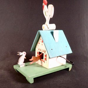May include: A wooden toy house with a blue roof and a white rooster on top. A brown rabbit is popping out of a window in the side of the house. A white rabbit is standing on a platform in front of the house.