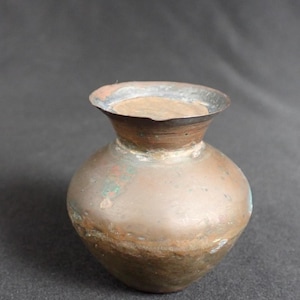 May include: A small, copper vase with a rounded body and a flared rim. The vase has a rustic, aged appearance with a greenish patina.
