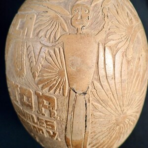 May include: A carved gourd with a human figure and intricate designs. The gourd is light brown and has a smooth surface. The figure is carved in relief and has a simple face with two eyes, a nose, and a mouth.