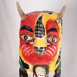May include: A wooden mask with horns, painted in red, yellow, black, and white. The mask features a fierce expression with large eyes and a wide mouth. The mask is decorated with intricate patterns and designs.