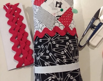May include: A black and white pincushion with a red and white polka dot fabric panel. The pincushion is filled with pins and safety pins. The pincushion is sitting on a white surface.