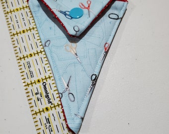 Op de afbeelding: Een kleine, driehoekige stoffen tas met een rode rand en een blauwe knoop. De tas is gemaakt van lichtblauwe stof met een patroon van scharen en naaiwerktuigen. De tas is ongeveer 12,5 cm lang.