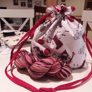 Puede incluir: Una bolsa de cordón roja y blanca con un estampado de ovejas y tejido. La bolsa está llena de un ovillo de hilo marrón, rosa y blanco.