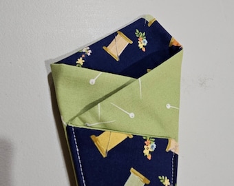 Op de afbeelding: Een groene en blauwe stoffen tas met een naai-thema. De tas is dubbelgevouwen en de binnenkant is zichtbaar. De stof heeft een patroon van spoelen garen, naalden en bloemen.