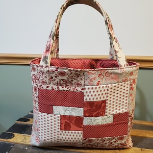 以下が含まれることがあります： 赤、白、クリーム色の生地で作られたパッチワークのトートバッグ。バッグは大きな長方形の形をしていて、布製のハンドルが2つ付いています。