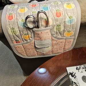Puede incluir: Un organizador de reposabrazos acolchado gris con un bolsillo para tijeras y un alfiletero. El organizador tiene un diseño de patchwork con alfileteros coloridos y el texto "Sewn with love".