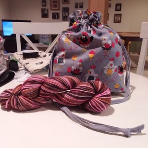 Könnte beinhalten: Eine graue Kordeltasche mit einem Hühnerdruckdesign. Die Tasche ist mit einem Knäuel rosa, braunen und weißen Garns gefüllt.