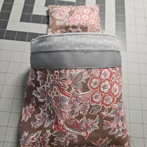May include: A brown fabric storage bin with a floral pattern in shades of red, blue, and white. The bin has a gray ribbon around the top and a white fabric lining with a small white cross pattern.