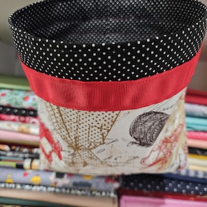 May include: A small, open-topped fabric basket with a black and white polka dot interior and handles. The exterior features a red ribbon and a cream-colored fabric with a vintage-style print. The basket is designed to hold small items.
