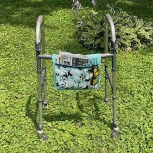 Op de afbeelding: Een zilveren rollator met een turquoise stoffen tas bevestigd aan het frame. De tas heeft een vlindermotief en bevat een krant.