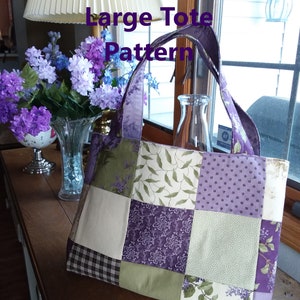 Op de afbeelding: Een grote tote bag gemaakt van patchwork stof in tinten paars, groen en crème. De tas heeft een vierkant ontwerp met verschillende patronen op elk vierkant. De tas is gelabeld "Lady In Stitches Large Tote Pattern".