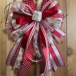 May include: A decorative bow made from multiple ribbons featuring a gingerbread man and cookie pattern, solid red satin ribbon, and red ribbon with white polka dots. The bow has several long, cascading tails and a central knot detail.