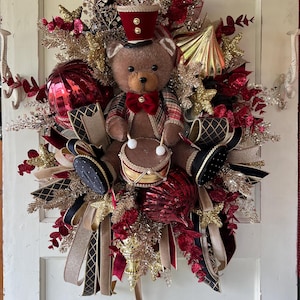 May include: A decorative holiday wreath featuring a central brown teddy bear wearing a red hat and plaid jacket, playing a small drum. The wreath is adorned with red and gold glittered foliage, large red and gold ornaments, and black and gold ribbons.