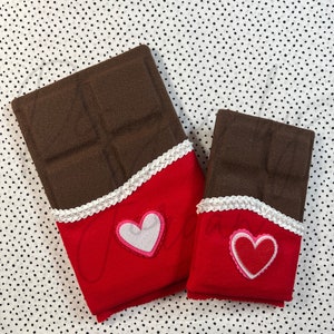 May include: Two chocolate bar shaped cushions with red fabric and white lace trim. The larger cushion has a pink heart with a white centre. The smaller cushion has a red heart with a white outline.