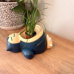 May include: A dark blue and cream-colored planter shaped like a reclining creature with closed eyes and small pointed ears. The planter features a circular opening on the back containing a small green plant with thin, grass-like leaves.