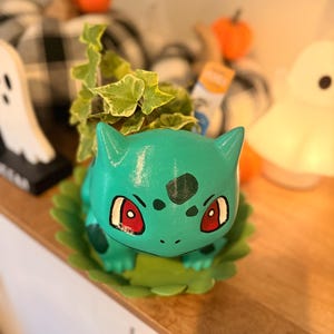 May include: A teal ceramic planter shaped like a fictional creature with large red eyes and dark green spots, featuring a green leaf-shaped base. A small variegated ivy plant grows from the top opening of the creature-shaped container.
