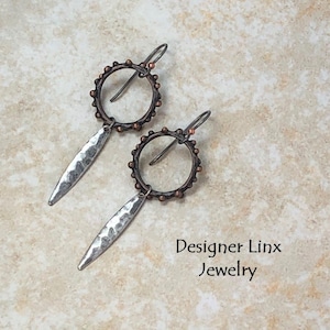 May include: A pair of silver and copper earrings with a hammered silver teardrop dangle and a copper circle with copper beads around the edge.  The earrings are on a light brown background.  Designer Linx Jewelry.