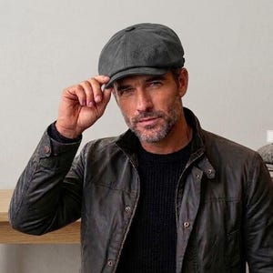 May include: A dark gray newsboy cap with a rounded crown and a short, stiff brim. The cap features a textured, matte finish and a central button detail on the top. The design is structured with multiple panels meeting at the center top button.