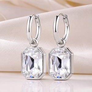 May include: A pair of silver-toned hoop earrings featuring rectangular, faceted clear stones hanging from each hoop. The earrings have a polished metallic finish and a classic design with a secure hinged closure for each circular hoop component.