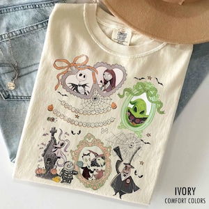 Puede incluir: Una camiseta de color marfil con un estampado gráfico de Pesadilla antes de Navidad. El diseño incluye a Jack Skellington y Sally en marcos con forma de corazón, Oogie Boogie en un marco verde, Zero y otros personajes con telarañas.