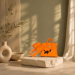 Könnte beinhalten: Eine leuchtend orangefarbene Handtasche mit goldfarbenen Beschlägen, einem Vorhängeschloss und einem kleinen schwarzen pferdeförmigen Anhänger, kombiniert mit passenden orangefarbenen Pantoletten mit H-förmigem Riemen auf einem Steinpodest.