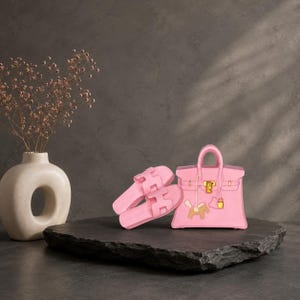 Pode incluir: Um par de sandálias de enfiar rosa claro com uma tira recortada em forma de H e uma mala de mão rosa claro a condizer com ferragens douradas, um fecho central, uma pega superior e um pequeno pendente em forma de cavalo.