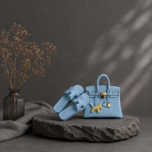 Pode incluir: Uma mala de mão azul-clara com ferragens douradas, um pequeno pendente de cavalo amarelo e um cadeado a condizer. Ao lado da mala estão duas sandálias de enfiar azul-claras com um recorte em forma de H na tira. Os itens são texturados.