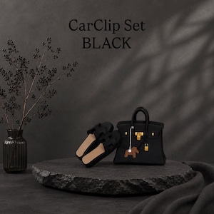 Pode incluir: Um CarClip Set BLACK composto por uma bolsa preta em miniatura com ferragens douradas e um pequeno pingente de cavalo castanho, acompanhado por um par de sandálias pretas com tira recortada em forma de H, sobre uma base de pedra.
