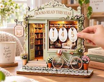 May include: A miniature wooden bookshop model featuring a sage green facade, a hanging sign reading Olivia's Bookshop, and a Books Read This Year header. It includes three oval number plaques, a bicycle, potted flowers, and an interior library scene.