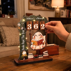 Peut inclure: Un présentoir à livres en bois représentant un lapin blanc portant des lunettes et lisant un livre. La structure comprend trois tuiles en bois suspendues affichant le nombre 362, le texte William's Library et Books Read This Year, éclairé par des guirlandes.
